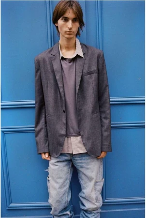 Young male model in grey blazer, striped shirt, and light denim jeans standing against a vibrant blue paneled door.