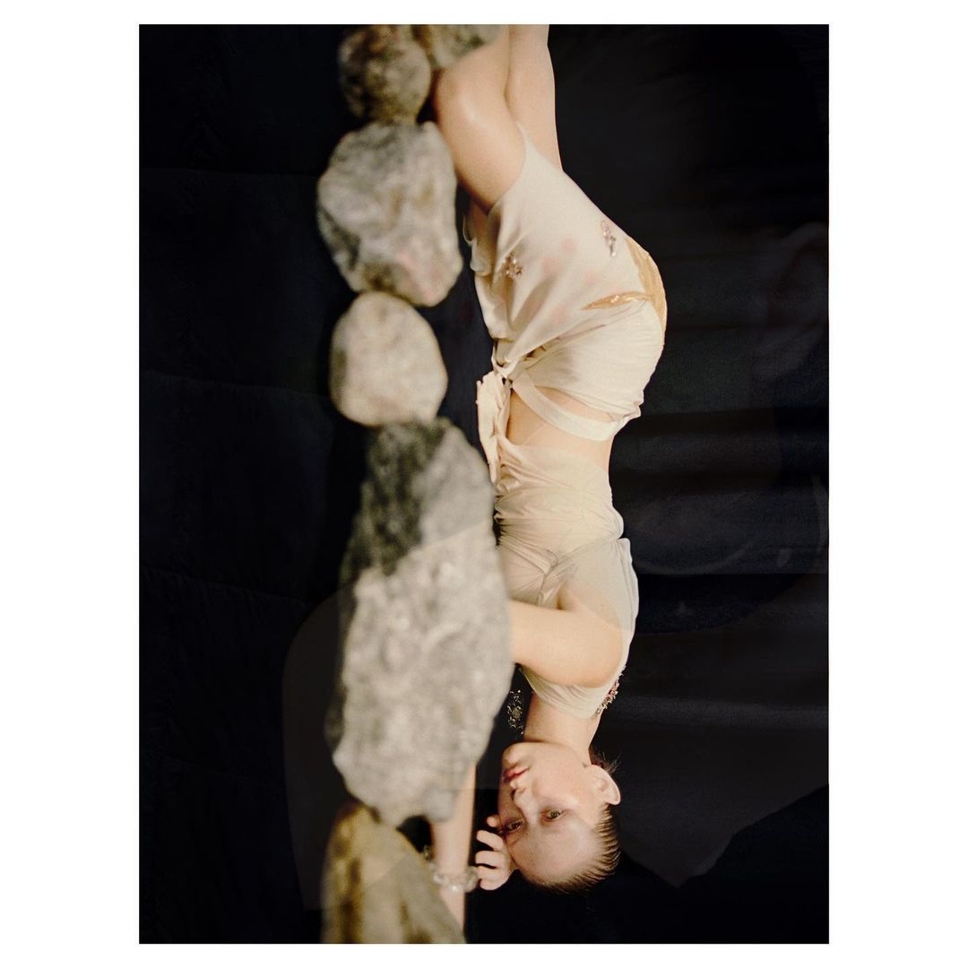 Upside-down studio shot of a woman in a draped garment alongside a stack of rocks against a dark background, serene.