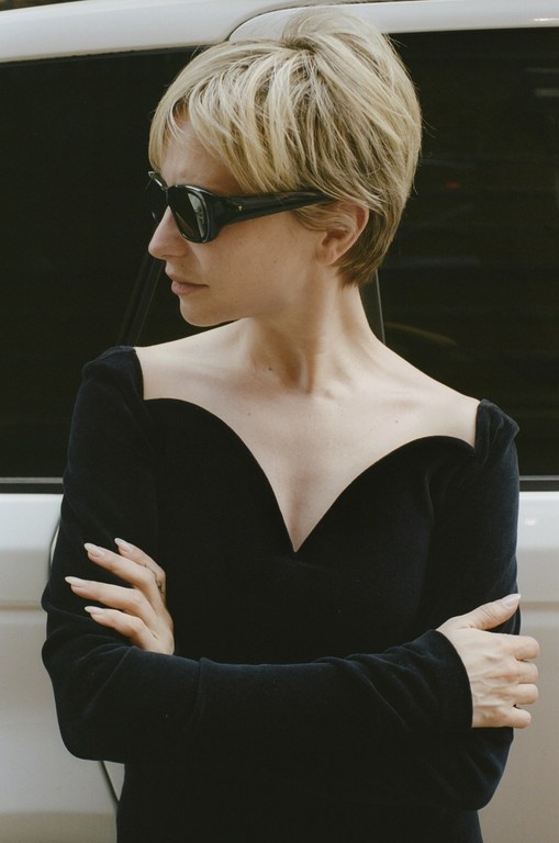 Portrait of a woman with short blonde hair and sunglasses wearing a black dress with a sweetheart neckline posing in front of a white car.