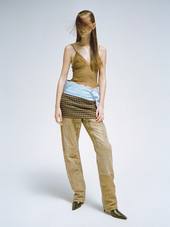 Full-length fashion shot of a slender model with long brown hair wearing a brown top, plaid skirt, and tan pants against a white backdrop.