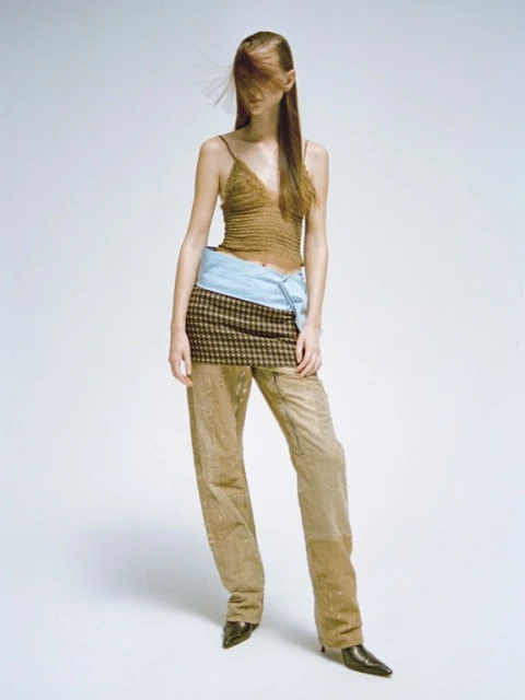 Full-length fashion shot of a slender model with long brown hair wearing a brown top, plaid skirt, and tan pants against a white backdrop.