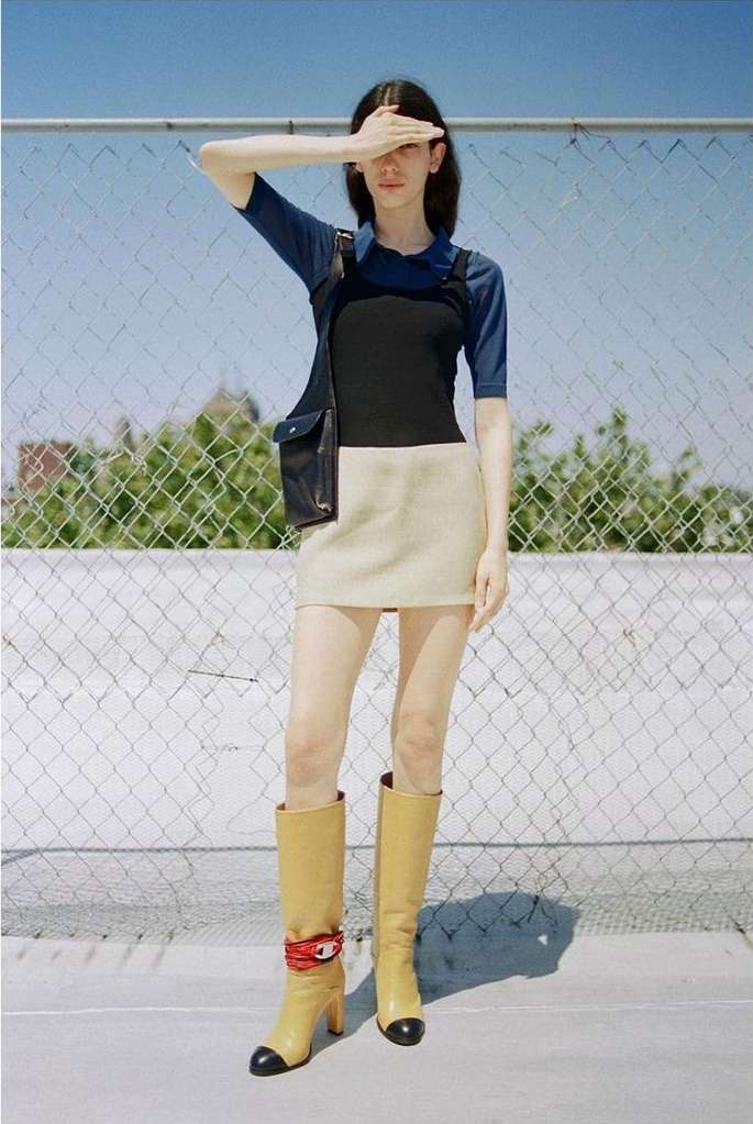 Woman in blue top, black overalls, beige skirt, and high boots poses against a chain-link fence under a bright sky in an outdoor shot.