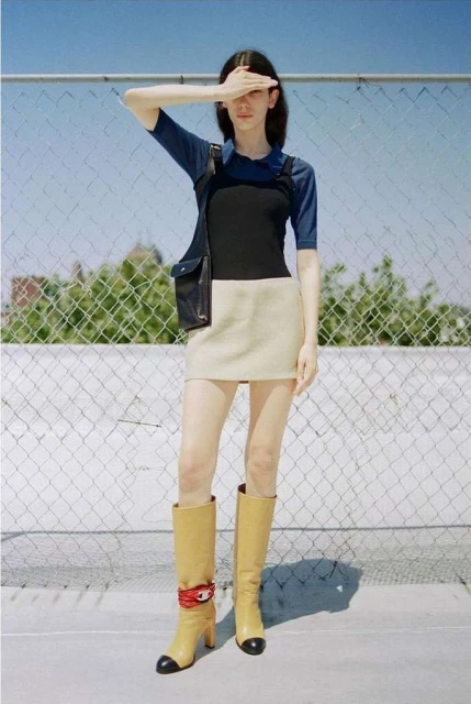 Woman in blue top, black overalls, beige skirt, and high boots poses against a chain-link fence under a bright sky in an outdoor shot.