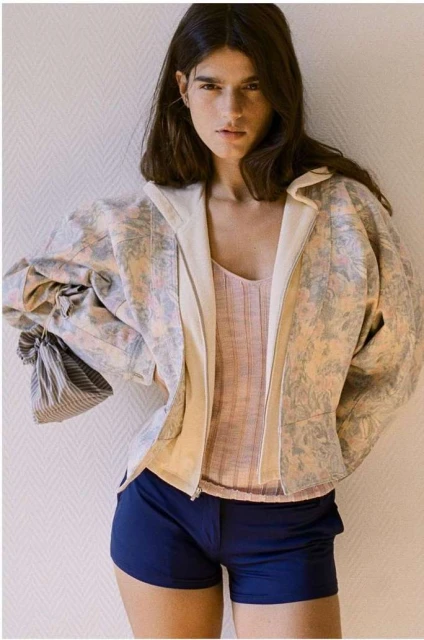 A woman in blue shorts and floral jacket poses indoors, showcasing fashion and style with a neutral background.