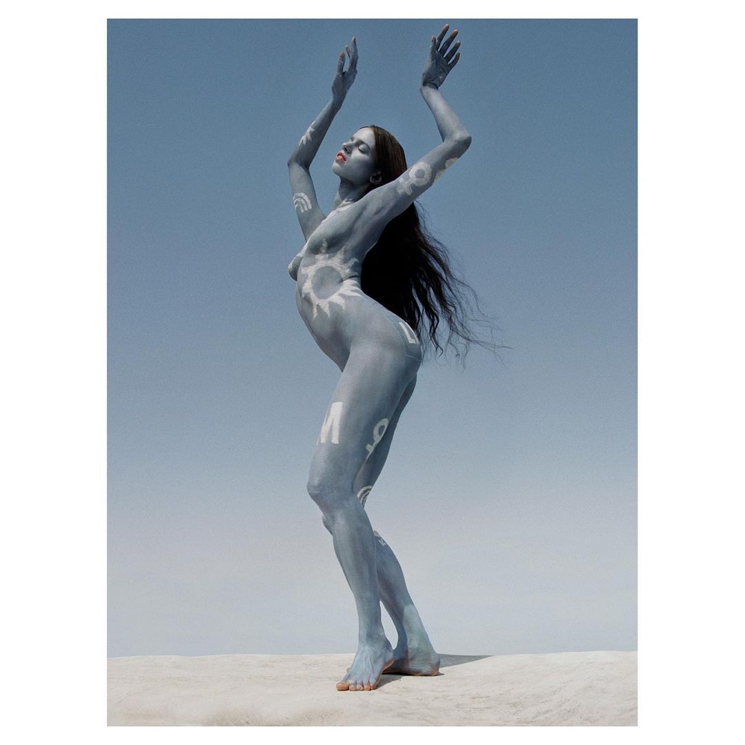 Artistic body painting on a nude woman with arms raised against a clear blue sky, standing on a neutral-toned surface.