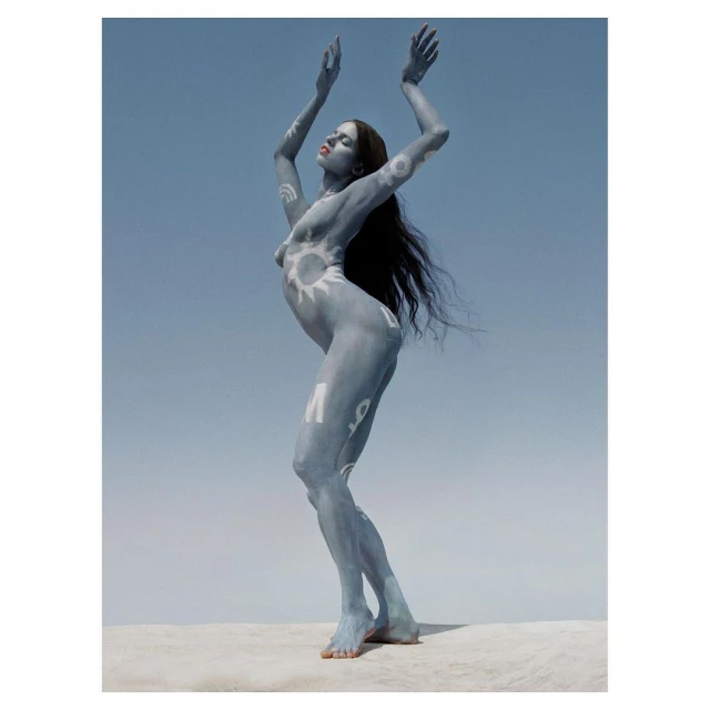 Artistic body painting on a nude woman with arms raised against a clear blue sky, standing on a neutral-toned surface.