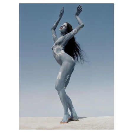 Artistic body painting on a nude woman with arms raised against a clear blue sky, standing on a neutral-toned surface.