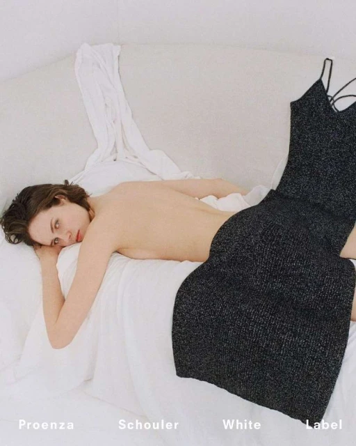 A woman lounges on a white bed next to a black glittery dress with delicate straps, with soft lighting and a bright, airy ambiance.