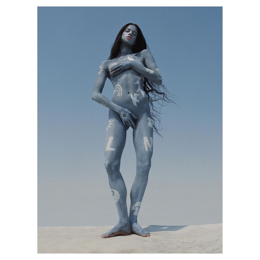 Nude female with body paint stands against a blue sky backdrop, creating a striking and artistic outdoor portrait with long hair.