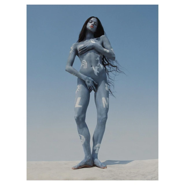 Nude female with body paint stands against a blue sky backdrop, creating a striking and artistic outdoor portrait with long hair.