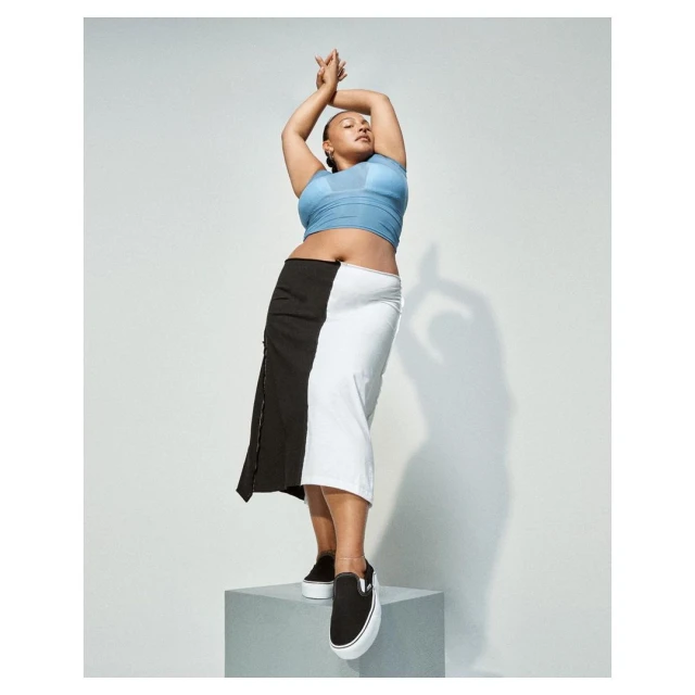 Full-length shot of a woman posing with arms raised, wearing a blue top, black and white skirt, and black shoes on a gray block.