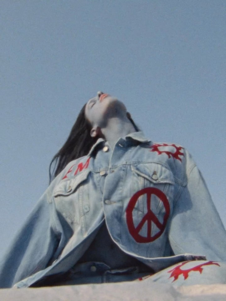 Woman with painted skin reclines in a denim jacket with a peace symbol against a clear sky, creating a retro, artistic vibe.