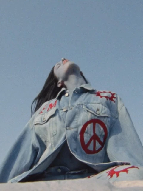 Woman with painted skin reclines in a denim jacket with a peace symbol against a clear sky, creating a retro, artistic vibe.