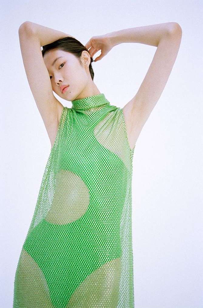 Woman in a sleeveless green mesh dress with hands above head against a white background, creating a fashion-forward and eye-catching image.