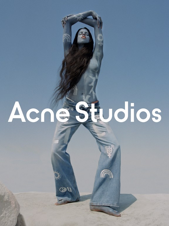 Fashion shot of a long-haired, blue-painted female model wearing denim pants with graphics, posing against a gradient blue sky backdrop.