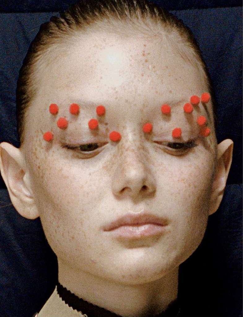 Close-up portrait of a fair-skinned adult female with red pom-poms above her eyebrows against a dark blue background.