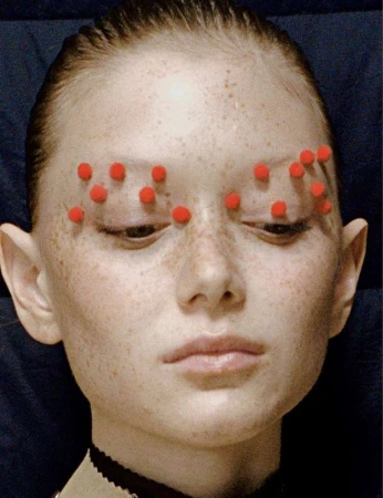 Close-up portrait of a fair-skinned adult female with red pom-poms above her eyebrows against a dark blue background.