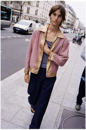 Man poses on a Paris street wearing a pink jacket with gold floral trim, a grey top, and dark pants, with buildings and vehicles in the background.