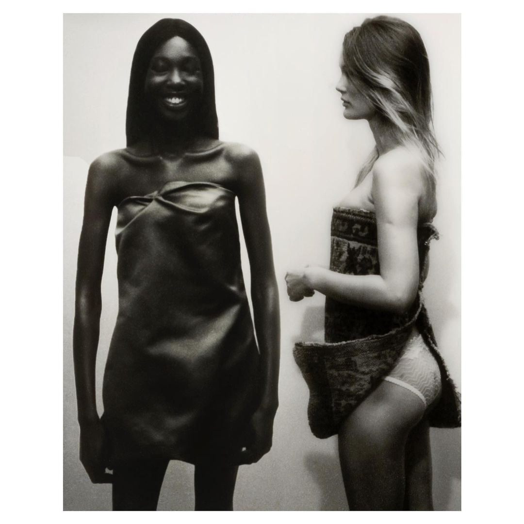 Monochrome shot of two fashion models in dresses, one with dark skin smiling, the other with light skin in profile, against a plain background.