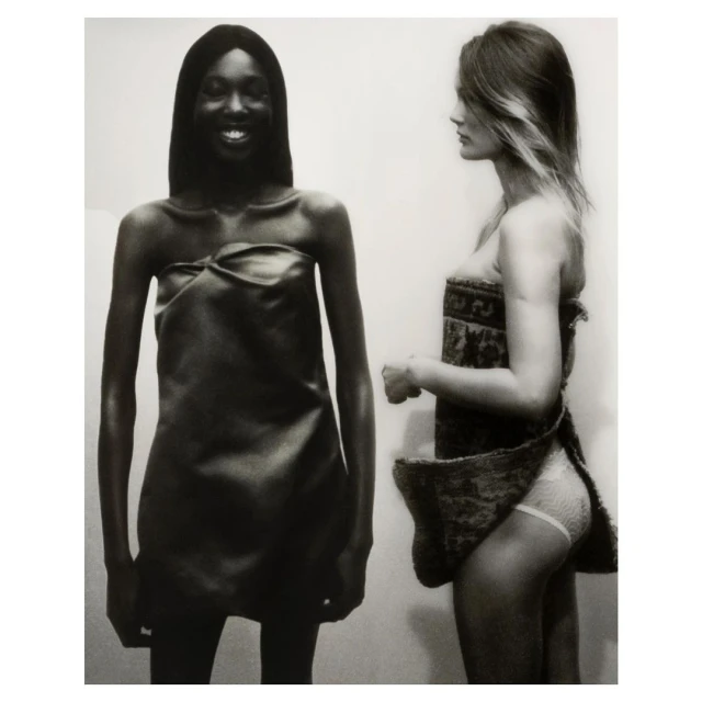 Monochrome shot of two fashion models in dresses, one with dark skin smiling, the other with light skin in profile, against a plain background.