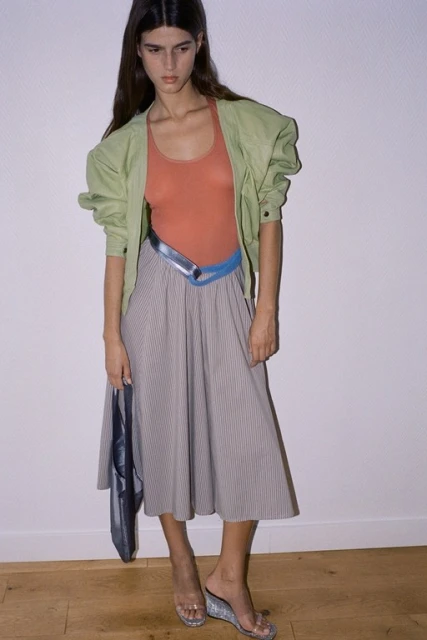 Fashion model poses in an indoor studio, wearing a light green jacket, orange top, striped grey skirt, and transparent heeled sandals.
