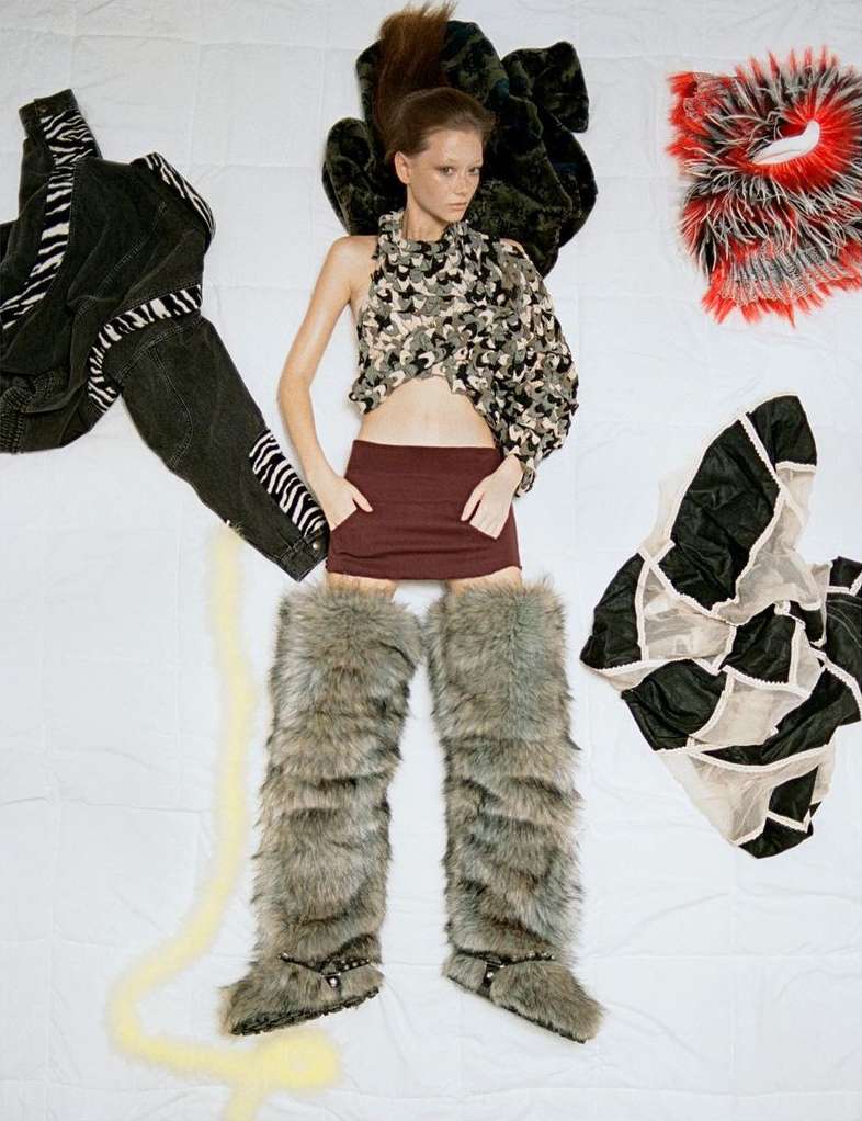 Fashion image: Young woman lies on white backdrop wearing an asymmetric top, burgundy skirt, and furry boots. Clothing items surround her.