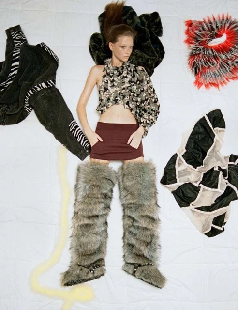 Fashion image: Young woman lies on white backdrop wearing an asymmetric top, burgundy skirt, and furry boots. Clothing items surround her.