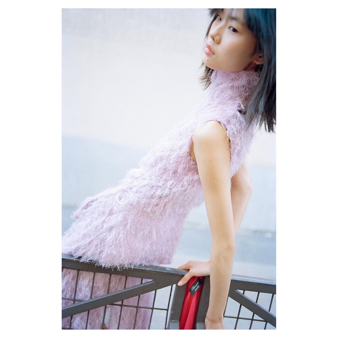 Asian woman poses in a sleeveless lavender dress with fuzzy texture, leaning against a dark railing with red fabric hanging.