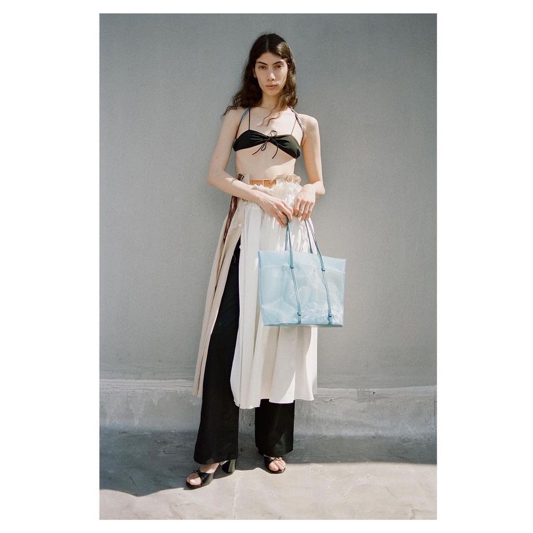 A tall adult woman poses outdoors in a black tie bikini top, high-waisted black pants, and a sheer white dress with a light-blue handbag.