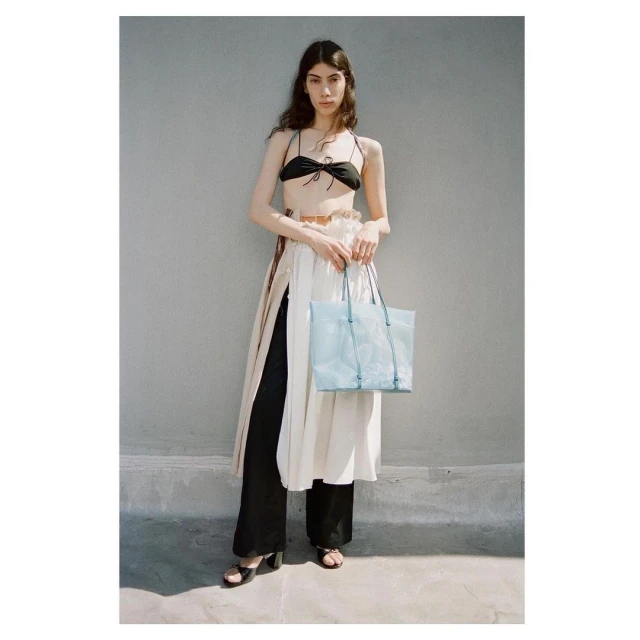 A tall adult woman poses outdoors in a black tie bikini top, high-waisted black pants, and a sheer white dress with a light-blue handbag.