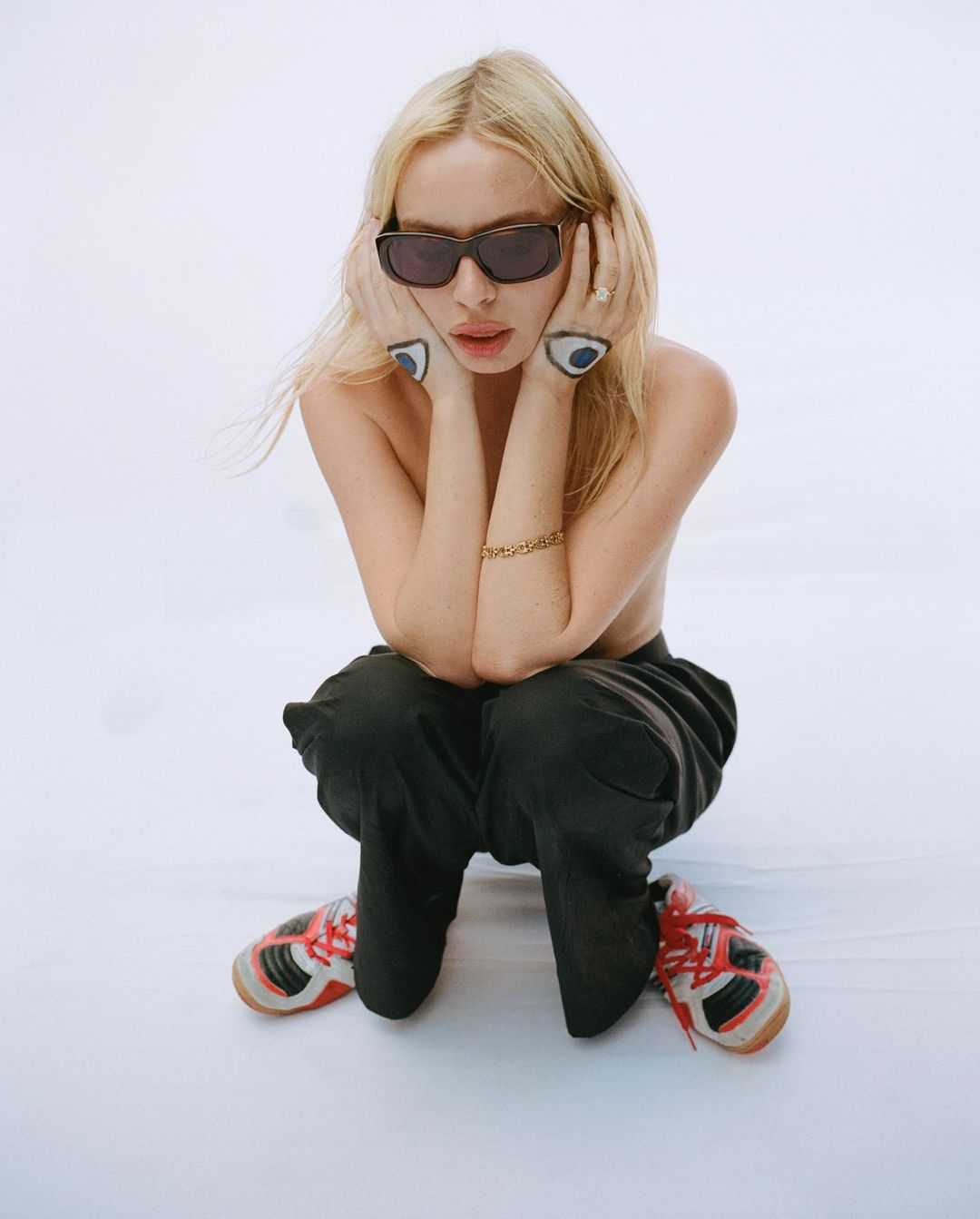 A woman with blonde hair in sunglasses squats in front of a white background, resting her hands on her cheeks. She is wearing sneakers.