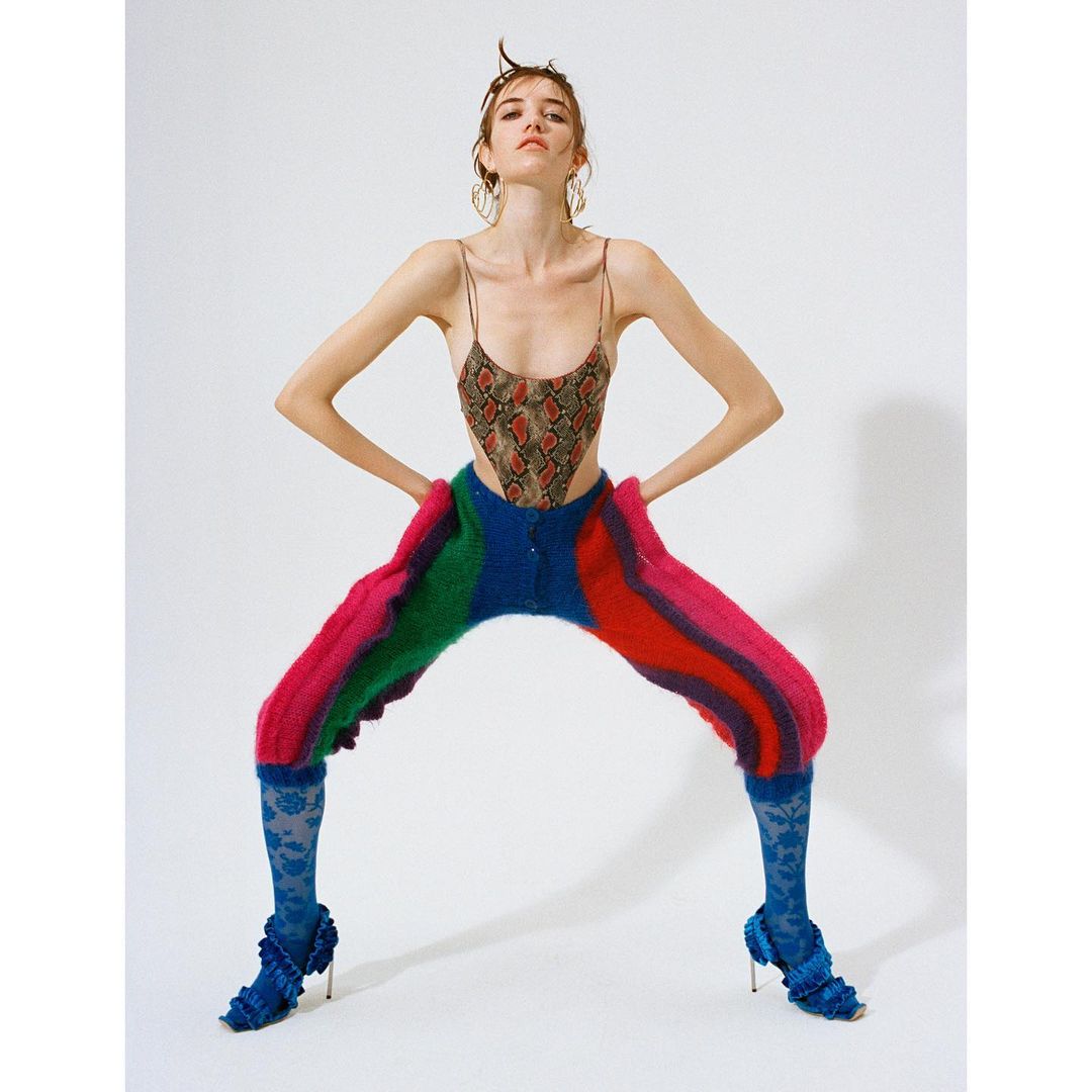 A fair-skinned woman in a snakeskin bodysuit, colorful knit pants, and blue floral-patterned heels poses against a white backdrop.
