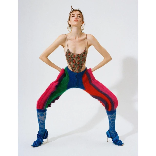 A fair-skinned woman in a snakeskin bodysuit, colorful knit pants, and blue floral-patterned heels poses against a white backdrop.
