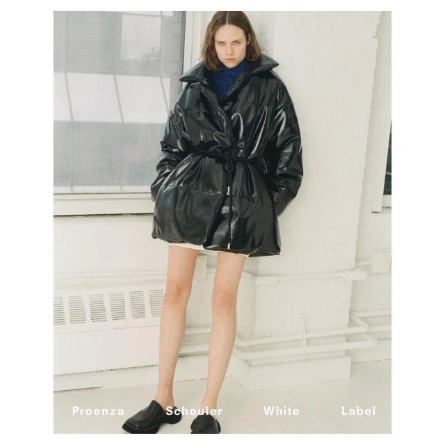 Adult woman posing in a glossy black puffer jacket and black flats in front of a white wall and window with light, neutral tones.