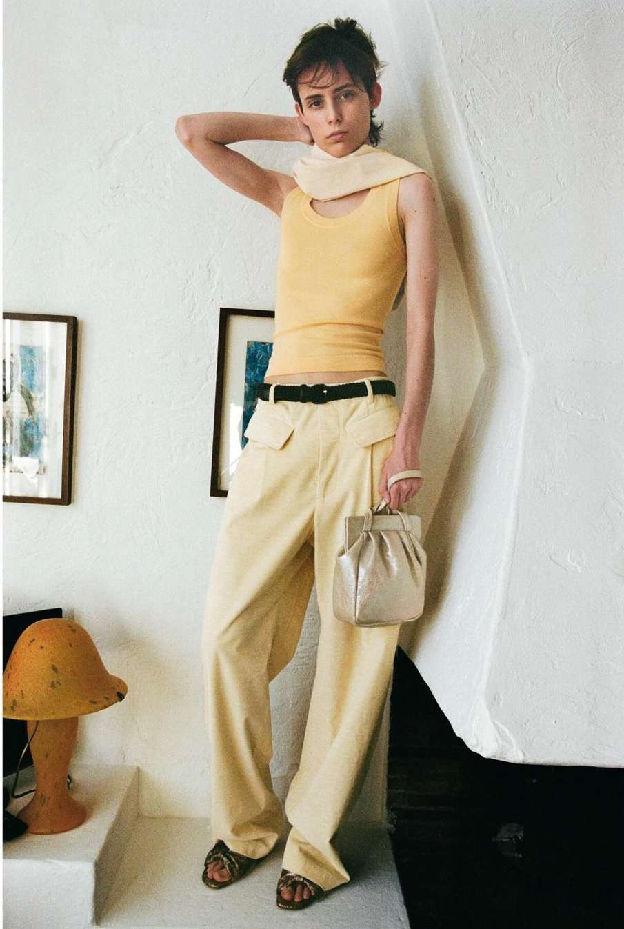 Fashion shot of a slender adult woman in wide-leg pants, tank top, and sandals, carrying a metallic purse in an indoor setting.