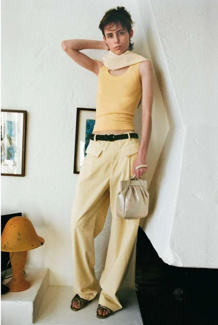 Fashion shot of a slender adult woman in wide-leg pants, tank top, and sandals, carrying a metallic purse in an indoor setting.