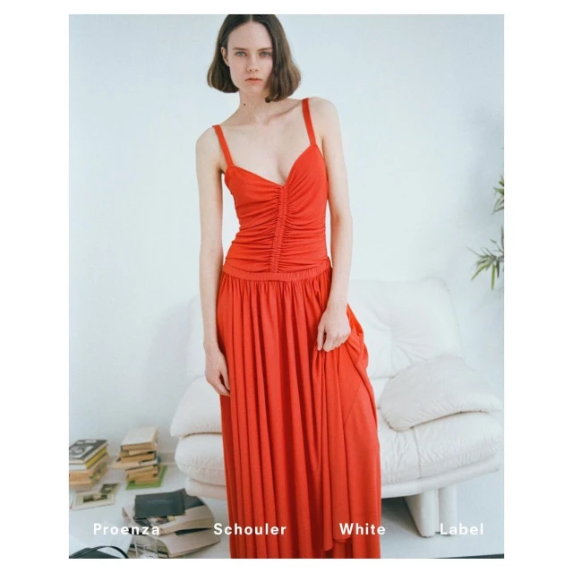 A slender adult female poses indoors against a white wall wearing a coral-red, floor-length dress, with a stack of books and a white sofa.