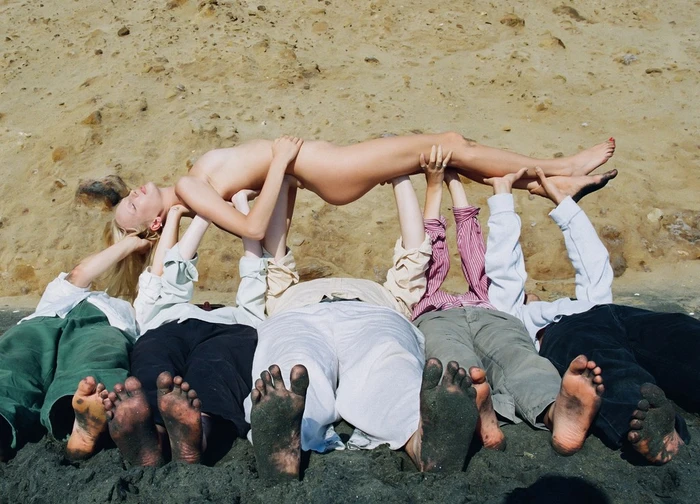 Group of people lying in dark sand holding up another person horizontally against a sandy slope.