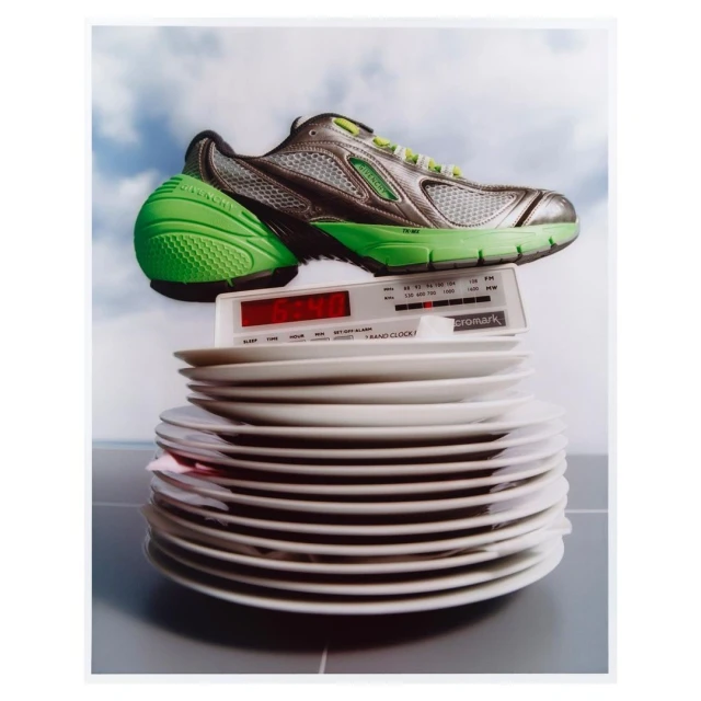 Still life featuring a running shoe on top of a vintage clock radio, which sits on a stack of plates against a cloud-filled sky.