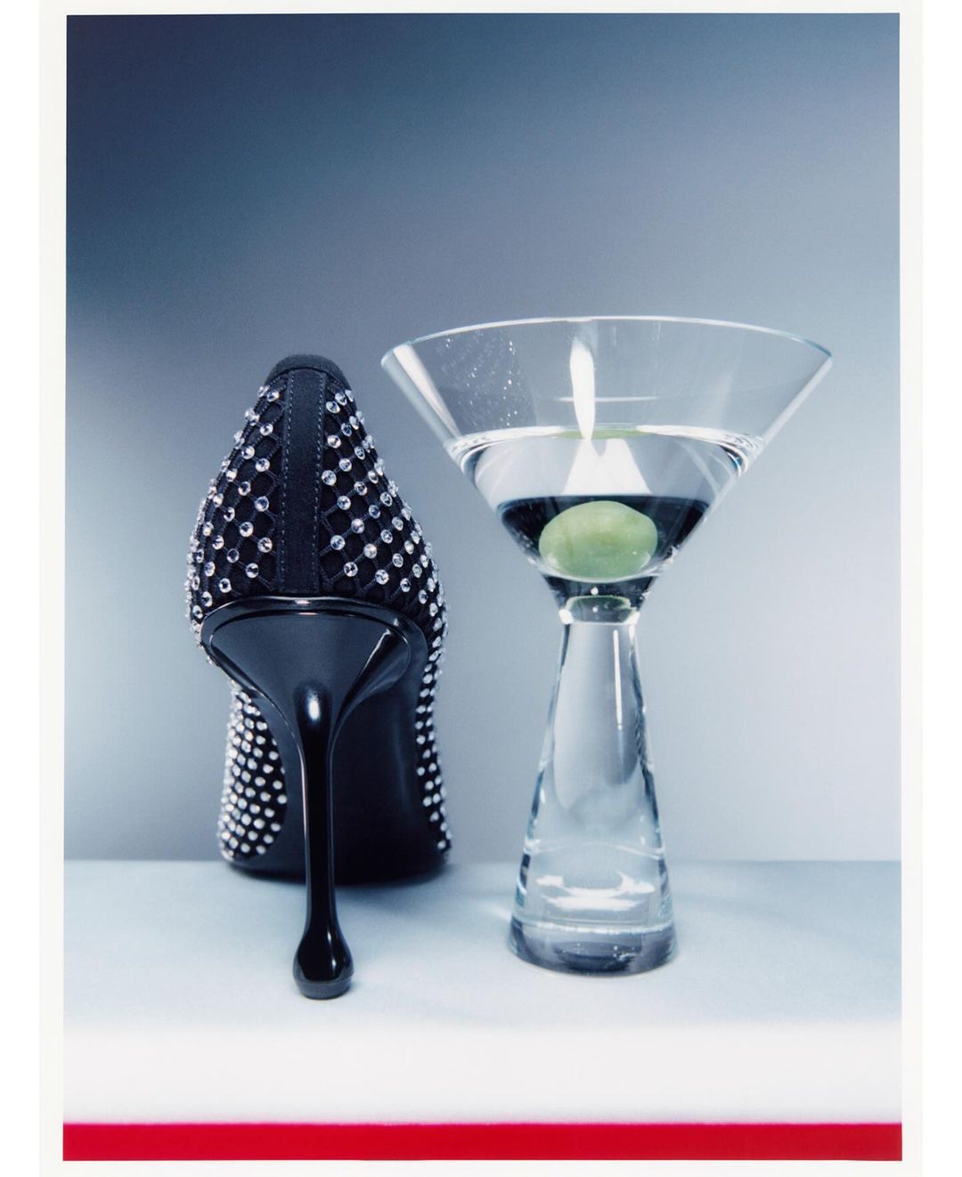 Crystal-embellished black high heel shoe standing beside a martini glass with an olive on a grey surface.