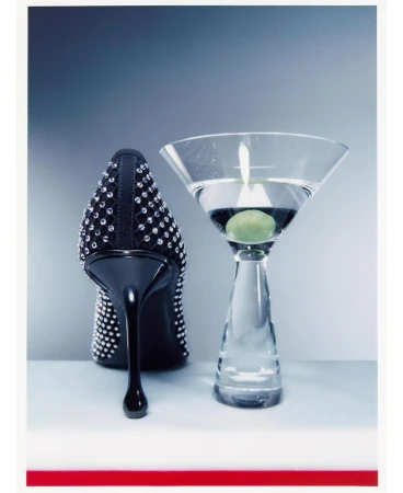 Crystal-embellished black high heel shoe standing beside a martini glass with an olive on a grey surface.