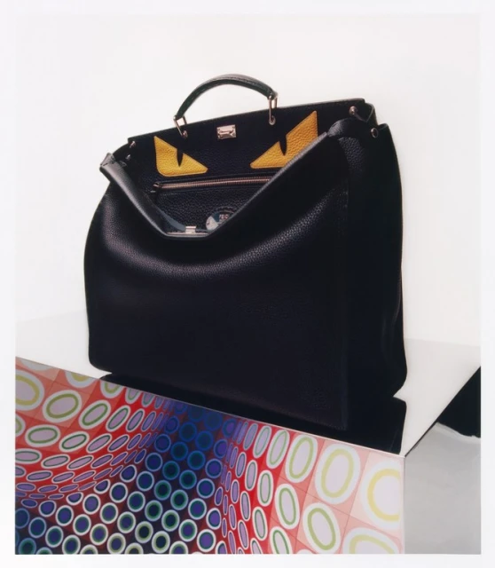 Black leather Fendi Peekaboo ISeeU East-West bag with yellow eye design displayed on a geometric stand against a white background.