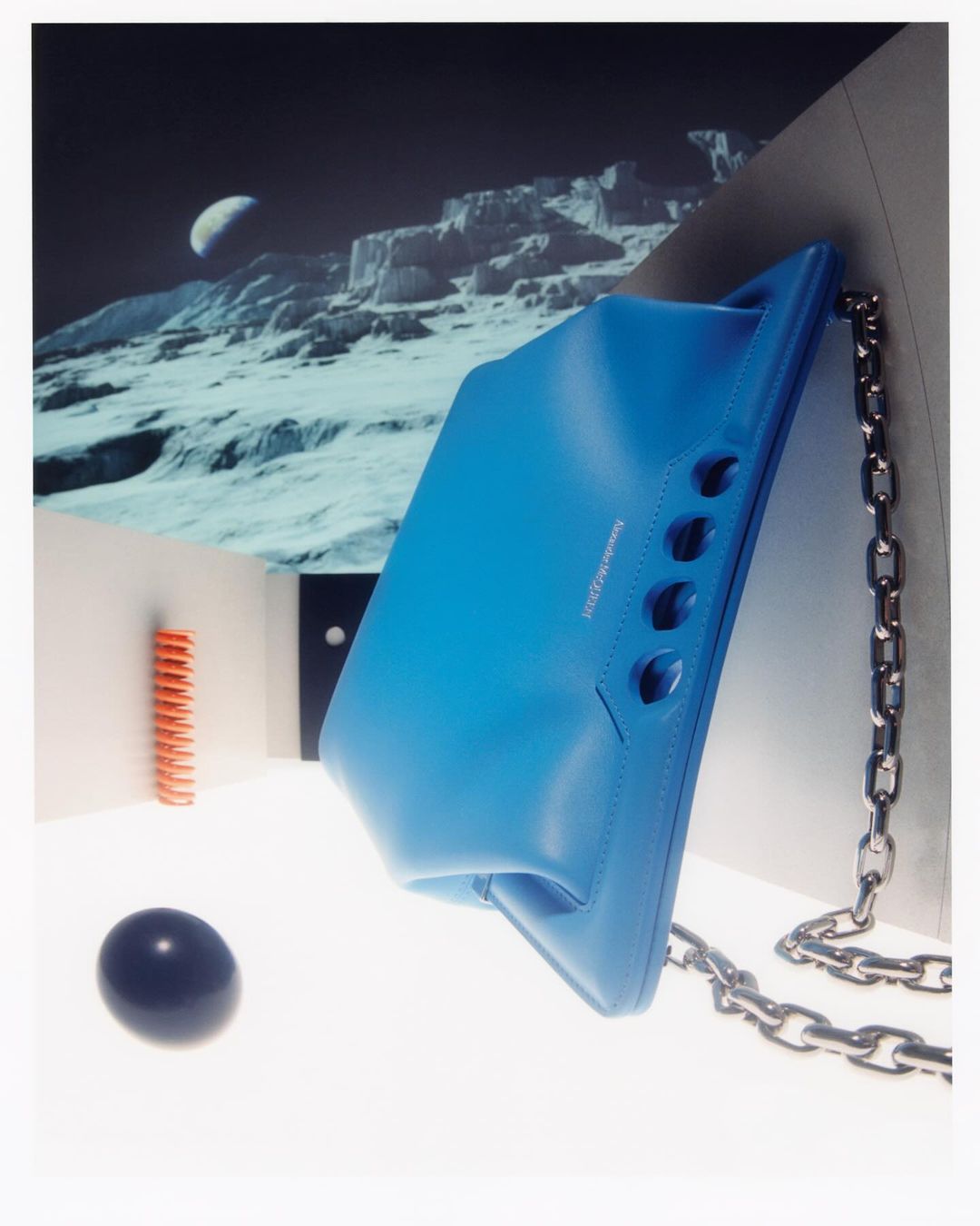 Eye-level studio shot of a blue leather handbag with a chain strap, contrasted against a white backdrop with a lunar landscape and black sphere.