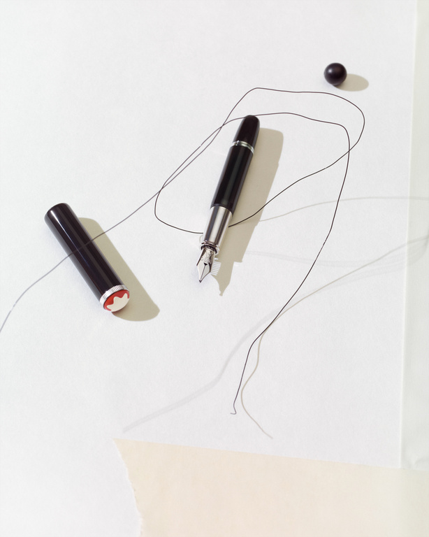 Elegant composition featuring a black fountain pen, its cap, a black sphere, and delicate black threads on a bright white surface.