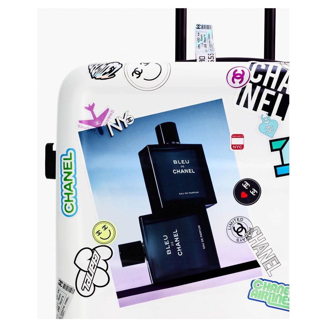 White suitcase featuring a Chanel Bleu de Chanel perfume advertisement and various Chanel-themed stickers and graphics in studio lighting.