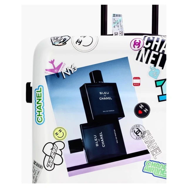 White suitcase featuring a Chanel Bleu de Chanel perfume advertisement and various Chanel-themed stickers and graphics in studio lighting.