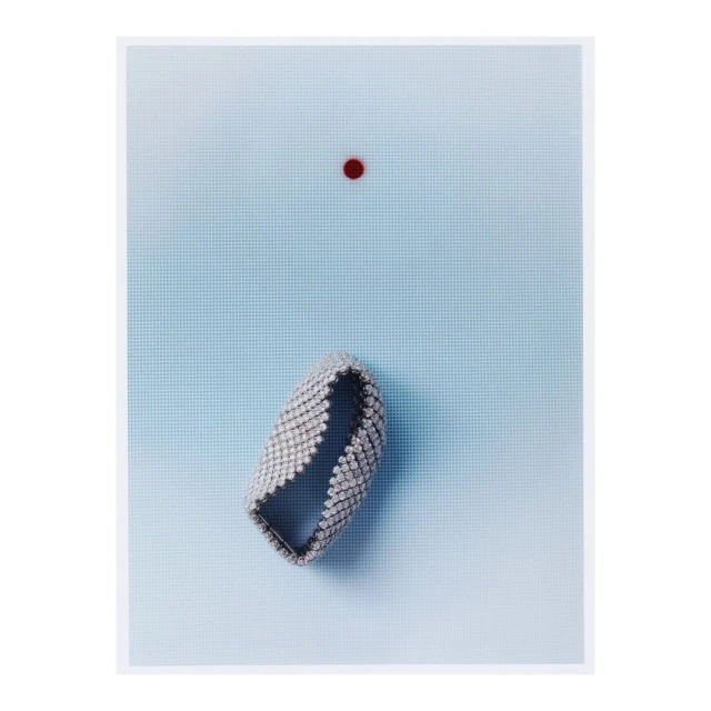 Elevated view of an open metal mesh bracelet on a light blue gradient background, accompanied by a single red dot above it.