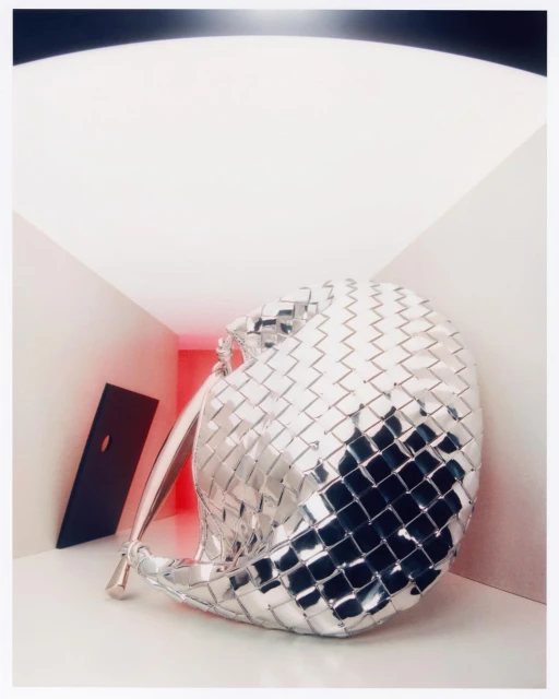 A silver metallic woven handbag is displayed in an indoor setting with bright, neutral lighting and a geometric black prop.