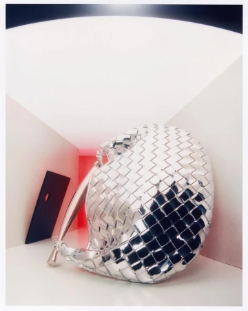 A silver metallic woven handbag is displayed in an indoor setting with bright, neutral lighting and a geometric black prop.