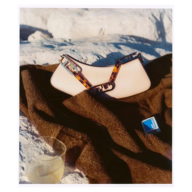 Fashionable purse with tortoise-shell chain on brown beach towel with drink and matchbox, against a blurred rocky background.